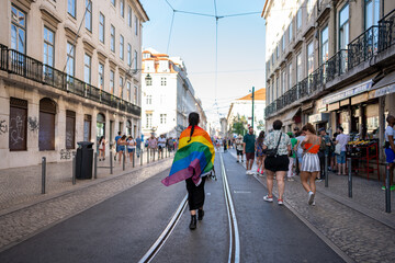 Back view of anonymous girl with rainbow flag gay pride parade walking on streets of Lisbon. LGBT community wearing rainbow flag strolling on crowded city street. Lesbian during pride month in