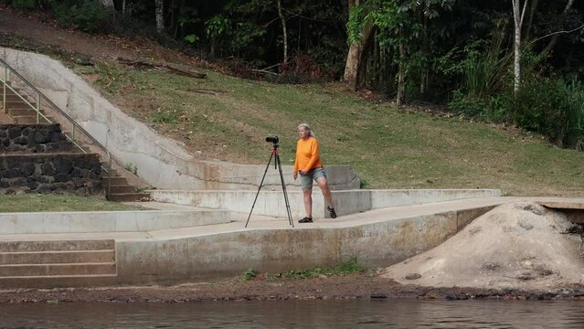 A woman in an orange jumper taking photographs with a camera on a tripod doing exercises as she waits for a long exposure photo to finish at Malanda falls on the Atherton Tablelands in tropical Queens