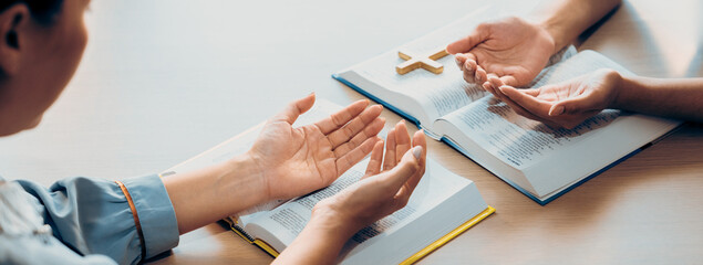Two believer praying together on holy bible book faithfully with wooden cross placed at wooden...