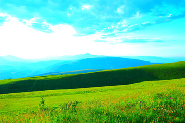 Sloping view in Piani di Ragnolo at green pastures punctuated by different beautiful flowers, a darker ridge with just a few trees, and the incredible blue outlines of the Sibillini in the background