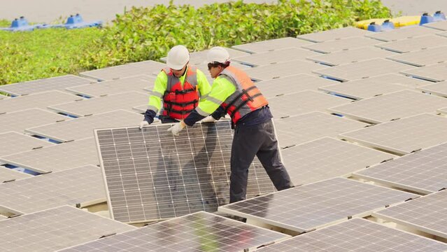 Two professional technician workers stand and hold solar cell panel in area of network of base over area of water reservoir.