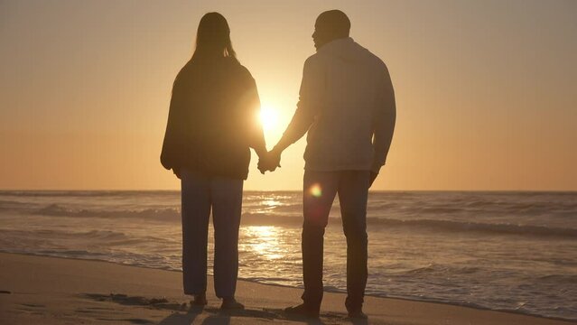 Camera pushes in on rear view shot of casually dressed loving young couple standing hand in hand Rear View Of Casually Dressed Loving Young Couple Standing Beach Shoreline Holding Hands At Sunrise
