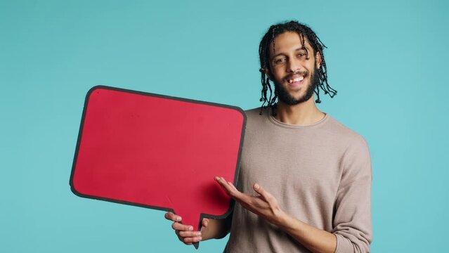 Happy man holding red speech bubble sign of empty space for text. Cheerful middle eastern person presenting thought bubble cardboard, isolated over blue studio background