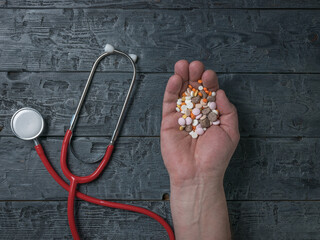 A man's hand with a lot of pills and a stethoscope on a wooden background.