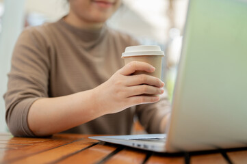 A woman holding a takeaway coffee cup, drinking coffee while working on her laptop at a cafe.