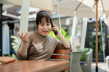 A positive woman is talking on the phone with someone while sitting at an outdoor table of a cafe.