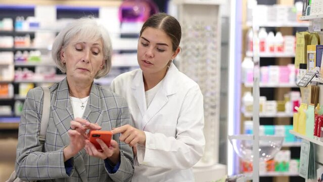 Pharmacist girl communicates with mature female client, looks at mobile phone screen, helps to identify desired drug in photo