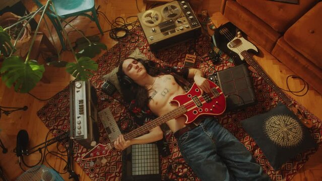 Young man in headphones and open shirt chilling on floor among audio recording devices, playing electric guitar and enjoying music. High angle view