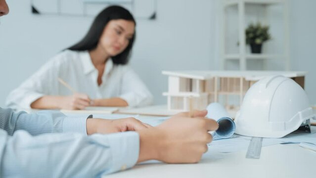 Professional architect engineer drawing blueprint and working together with young beautiful caucasian coworker on meeting table with safety helmet, house model, blueprint scatter around. Immaculate.