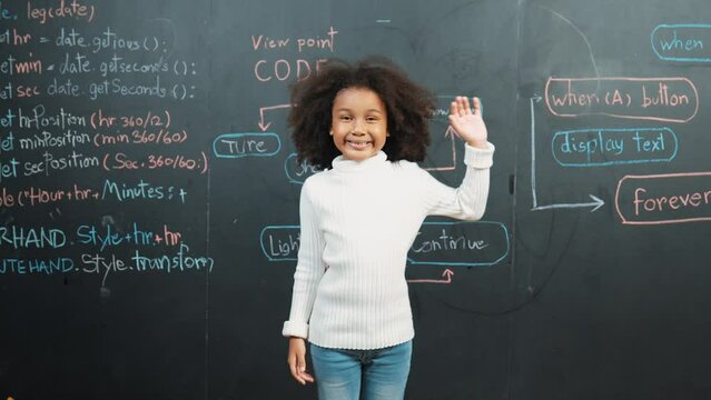 African girl smiling and waving at camera while standing at board with engineering prompt or coding, programing system written in STEM classroom. Child looking at camera while greeting. Erudition.