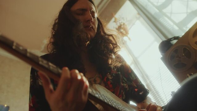 Young talented musician with curly long hair playing the guitar with eyes closed and enjoying melody in living room with sunlight shining through window. Rack focus, low angle shot
