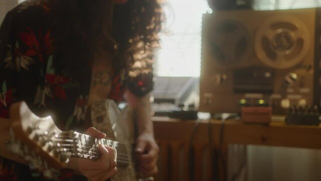Man with curly long hair playing the guitar beside vintage reel-to-reel audio tape recorder at home music studio. Midsection shot