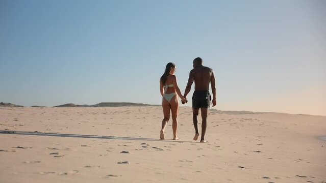 Camera tracks across rear view of loving young couple wearing swimwear holding hands walking along beach in South Africa - shot in slow motion