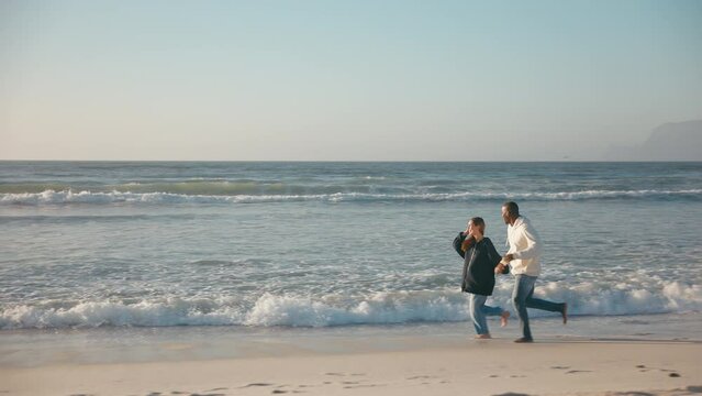 Camera tracks full length shot of casually dressed loving young couple running hand in hand along sandy shoreline of beach and sea in South Africa - shot in slow motion