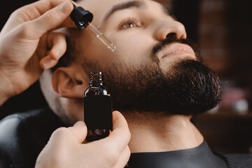 Barber applies beard oil with dropper for man in barbarshop.