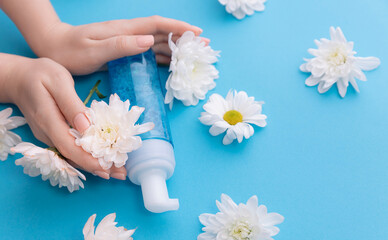 Flat lay woman hands with bottle with water foam for cleansing on blue background with white flowers. Concept banner organic cosmetics for facial skin care.