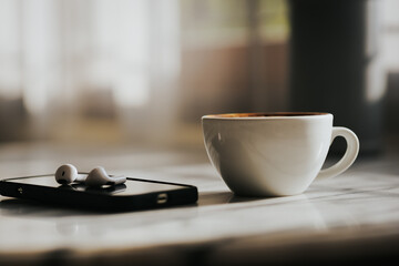 A cup of hot latte and a cell phone, headphones on a sunny wooden table by a window with white curtains. Vintage tones. Free space to create a message.