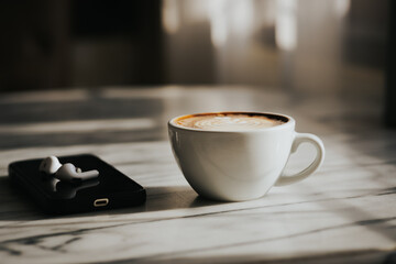A cup of hot latte and a cell phone, headphones on a sunny wooden table by a window with white curtains. Vintage tones. Free space to create a message.