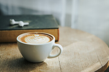 A cup of hot latte and a cell phone, headphones on a sunny wooden table by a window with white curtains. Vintage tones. Free space to create a message.