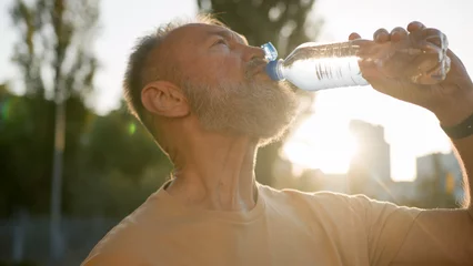 Gordijnen Close up older man drinking clear mineral water morning sun rays thirsty male drink pure aqua bottle city sunlight resting outside in park sport active sportsman healthy refreshing wellbeing energy © Yuliia
