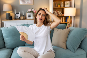 Obraz premium Woman puts head on sofa cushions feels sluggish due unbearable heat, waves hand fan cool herself, hot summer flat without air-conditioner climate control system concept