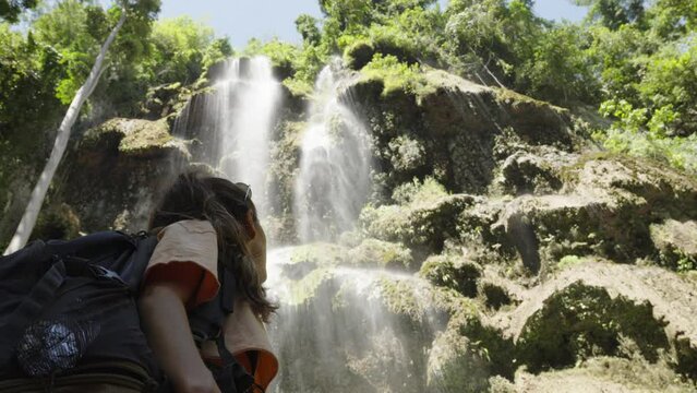 Young caucasian woman with adventuring backpack stares up at tropical Tumalog waterfall in Phillippines
