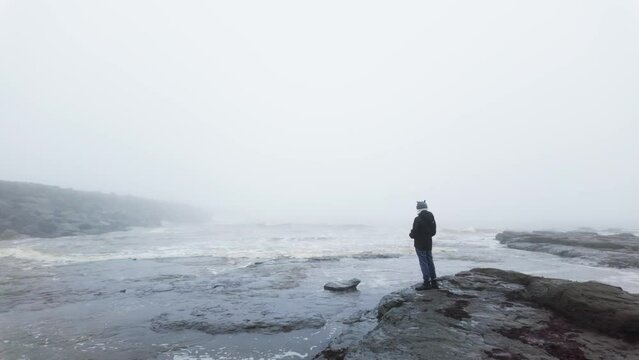 Young boy standing on the rocks on a coastal landscape. Winter scene with mist and fog. Seascape, outdoor clothing, crashing waves and rough seas