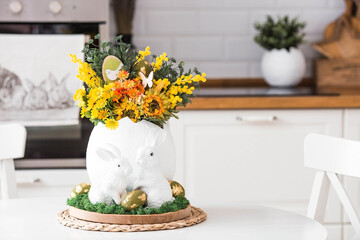 A spring bouquet in an egg, Easter bunnies and eggs with a golden pattern on the table. In the background is a white Scandinavian-style kitchen. A beautiful greeting card. Easter decor in the house.