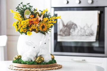 A spring bouquet in an egg, Easter bunnies and eggs with a golden pattern on the table. In the background is a white Scandinavian-style kitchen. A beautiful greeting card. Easter decor in the house.