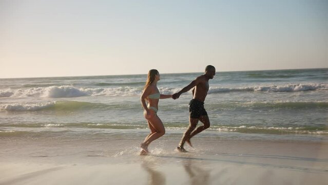 Camera tracks full length shot of loving young couple in swimwear holding hands and running along beach in South Africa - shot in slow motion