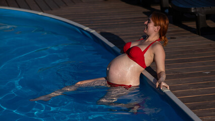 A red-haired pregnant woman in a red bikini is resting in the pool, leaning on the side.