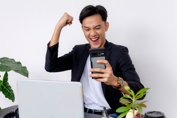 Excited young Asian man in a suit cheers on while looking at his smartphone, expressing joy at...