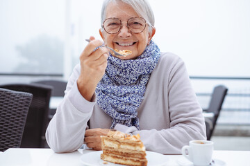 Happy senior woman sitting at the cafe table with a slice of coffee cake. Elderly woman ignoring the diet, committing a sin of gluttony - obrazy, fototapety, plakaty