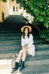 a cute girl in a hat is sitting on the stairs of the old town. Brunette in a white dress walks the street - 745590258