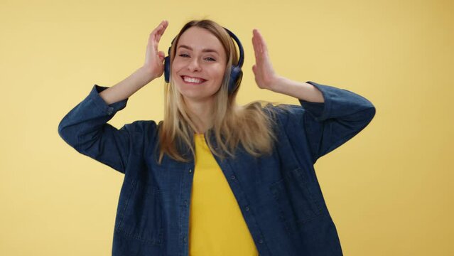 Energetic young female actively moving while listening music in dark blue wireless headset indoors. Joyful blond caucasian woman enjoying zippy tracks over isolated yellow studio background.