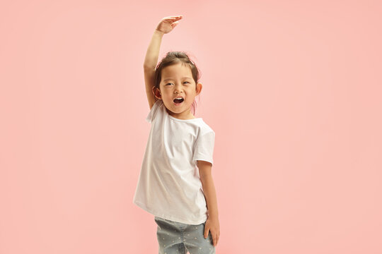 Cheerful Asian ethnicity little girl playfully stretches arm upwards, showing her height, denoting how she grew up, reaching for the sky, symbolizing growth, ambition, standing on pink isolated wall