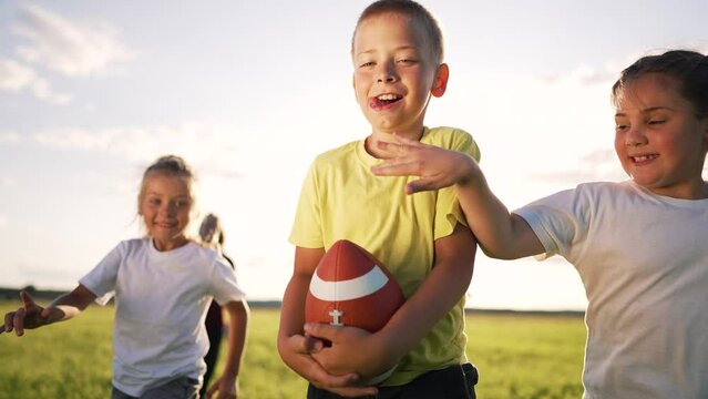 Happy team of children in park at sunset. Children play rugby ball in american football in park. Summer games children in park in nature run on the green grass. Family plays runs in team games in park