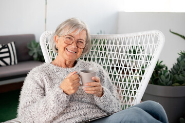 Portrait of senior attractive woman with cheerful smile relaxing at the armchair in the balcony at home looking at camera holding a coffee cup
