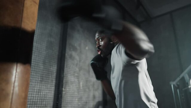 Boxer training hitting punching bag. African american sweated sportsman practicing punches in gym wearing boxing gloves. Workout, martial art, training, sport, kickboxing, boxing practice concept.