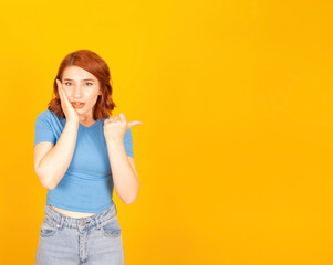Shocked young caucasian red bob hair woman pointing aside copy space with thumb. Touching her cheek with hand, open mouth. Yellow studio background. Demonstrating workspace area.