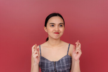 Portrait of an optimistic young Filipino woman crossing her fingers against a red background, symbolizing hope and wish.