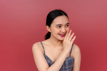 A young Asian woman whispers with a smile, hand on mouth, signifying secrecy or gossip against a plain background. - obrazy, fototapety, plakaty