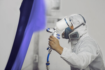 Male worker in protective clothes and mask painting part of car to blue violet color using spray...