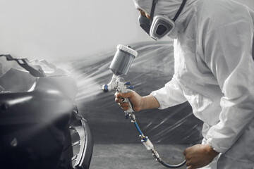 Spray painter worker in protective glove with airbrush pulverizer varnishing and painting part of car body in paint chamber.