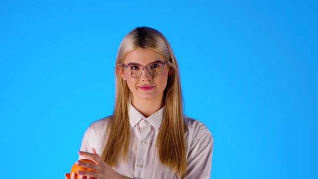 Smart looking blonde caucasian young woman with glasses plays with an orange happy, infinite background chroma blue portrait shot