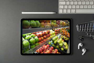 Tablet with grocery section on screen on dark gray office desk. There is a grocery cart and a keyboard next to him