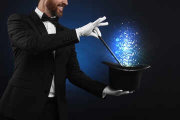Magician showing trick with wand and top hat on dark background, closeup. Fantastic light coming...