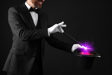 Magician showing trick with wand and top hat on dark background, closeup. Fantastic light coming...
