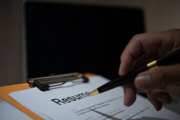Hand holding a pen over a resume on a clipboard with a dimly lit background.