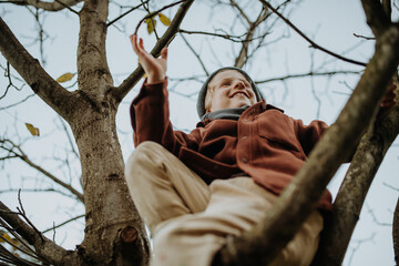 Happy boy playing on tree during first spring days. Kid climbing tree, sitting on branch and smiling. Outdoor, garden activity for young children. - 745641830